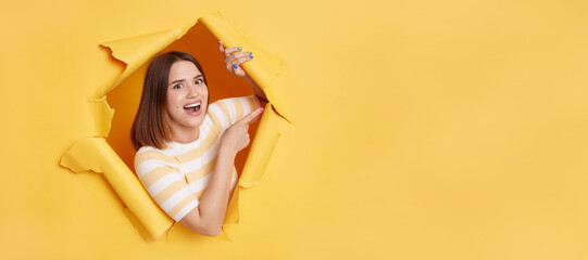 Indoor shot of amazed young attractive woman wearing striped T-shirt breaking through hole in...