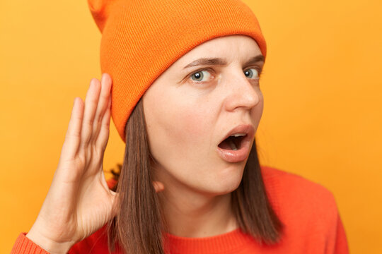 Portrait of shocked hipster woman wearing orange sweater and hat, keeps hand near ear, hearing astonishing talking, posing isolated over yellow background.
