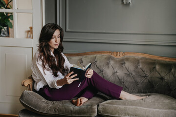 Exhausted brunette businesswoman with dar wavy hair sitting on sofa wearing white t-shirt and violet pants holding diary and leaning on hand in upset expression. Overloaded woman, loosing job.