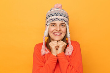 Portrait of cute delighted Caucasian woman wearing funny earflap hat and orange casual jumper, looking at camera, keeps fists under chin, standing isolated over yellow background.