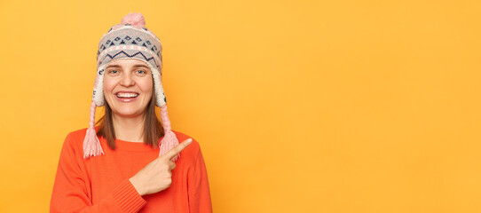 Photo of smiling attractive woman wearing funny earflap hat and orange casual jumper standing isolated over yellow background, pointing at advertisement area, mockup for promotion.