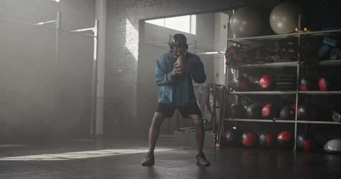 Sporty mad furious African male successful rugby player in blue uniform and helmet aggressively playing with ball screaming in dark authentic gym. Angry American football athlete preparing for match.