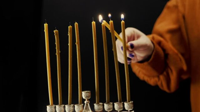 A woman's hand lights candles on the last day of Hanukkah from the shamash candle in the Hanukkah menorah. Medium plan