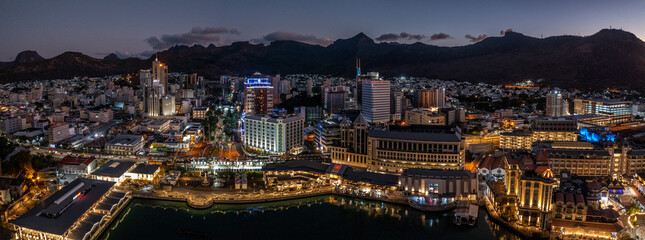 panoramic aerial night view of the old and modern buildings at Waterfront in Port Louis, Capital of Mauritius with mountains in the background. Bird eyes view during nighttime  - obrazy, fototapety, plakaty
