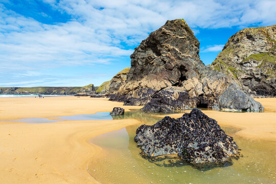 Sandy beach with rocky formations along a rocky cliff shoreline with surf and blue sky and clouds; Cornwall County, England