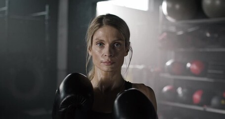 Close-up portrait of blonde young professional woman boxer preparing for battle wearing gloves and top in dark gym. Attractive angry focused confident female boxing athlete looking at camera.