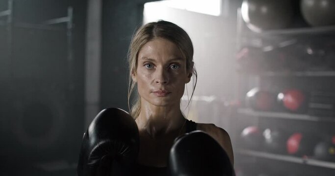 Close-up portrait of blonde young professional woman boxer preparing for battle wearing gloves and top in dark gym. Attractive angry focused confident female boxing athlete looking at camera.