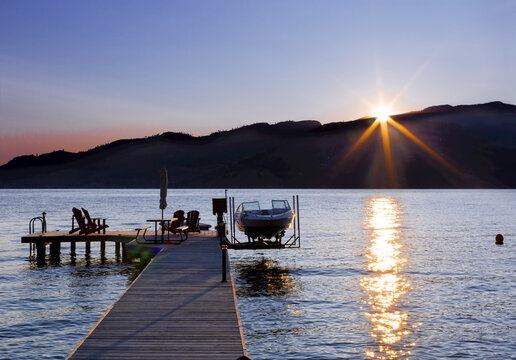 Adirondack Chairs And A Picnic Table Sit On A Wooden Dock With A Boat And Lift On Okanagan Lake At Sunset; British Columbia, Canada