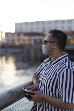 A man wearing a face mask and sunglasses stands looking out and using his smart phone, Lonsdale Quay; North Vancouver, British Columbia, Canada