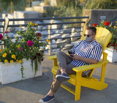 A man wearing a face mask and using his smart phone sits in a chair at Lonsdale Quay; North Vancouver, British Columbia, Canada