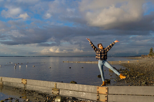 A young woman wearing a face mask stands in a silly pose on a narrow beam on Crescent Beach along the coast of BC; Surrey, British Columbia, Canada
