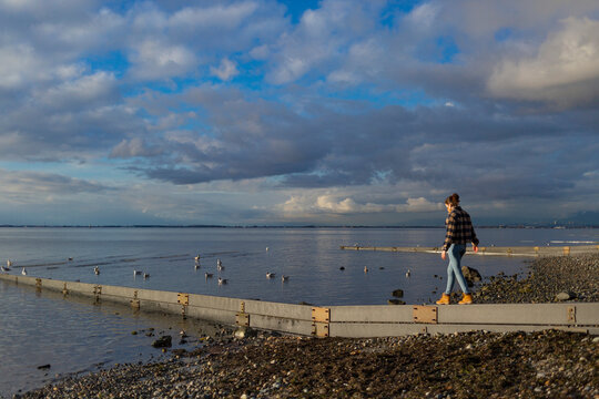 A young woman wearing a face mask walks along a narrow beam at the water's edge on Crescent Beach along the coast of BC; Surrey, British Columbia, Canada