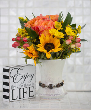 A motivational message on a wooden block 'Enjoy the little things in life' with a beautiful and colourful bouquet of fresh cut flowers on a white tile surface; Studio