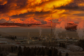 Dramatically lite gas plant at sunrise with glowing snow covered mountain range in the background with glowing clouds in the sky, West of Calgary; Alberta, Canada