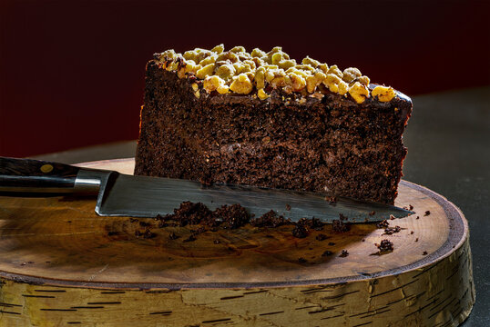A cut piece of chocolate cake topped with nuts on a cut tree trunk with a knife; Studio