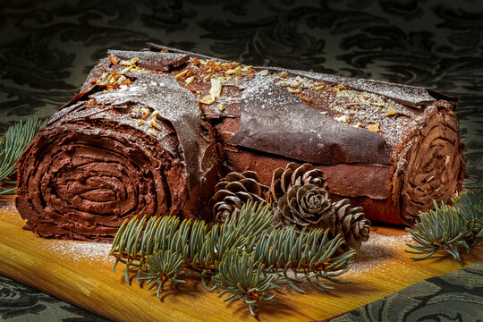 Icing frosted rolled chocolate Yule log cake with evergreen tree pieces and pine cones on a wooden board sitting on a dark green decorative table cloth; Calgary, Alberta, Canada