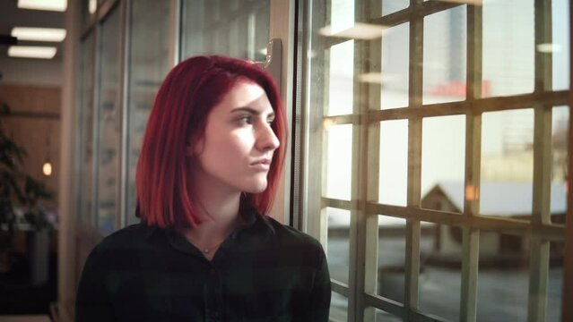 Young developer woman with red hair lost in thoughts looking away through the office window. Businesswoman planning or trying to solve some business issues