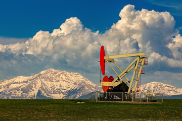 Pumpjack in a green field with dramatic storm clouds, snow-covered mountain range and blue sky in the distance, North of Longview, Alberta; Alberta, Canada