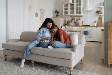 Depressed unhappy teenage girl experiencing stress after getting bad grades at school or quarrel with classmates. Loving caring woman calms upset child hugging daughter sits on sofa in apartment