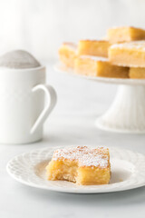 A delicious lemon bar with a bite out of it and a stand of bars in behind.