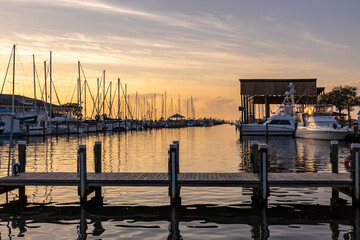 An inlet at sunset with sailboats, a boat house, yachts and a pier across the bottom of the scene. - Powered by Adobe