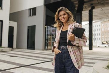 Pretty young caucasian business woman holding tablet posing in front of city building. Curly blonde wears jacket and jeans. People, lifestyle and communication concept.