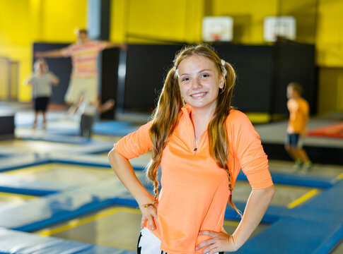 Portrait Of Positive Smiling Teen Girl In Orange Jumper Posing On Trampolines During Weekend Free Time At Game Center