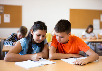 Girl and boy sitting at desk in classroom with their classmates, listening to teacher explaining...