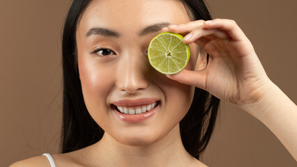 Fruit vitamins for beauty. Young asian woman holding lime in front of her eye, posing over brown...