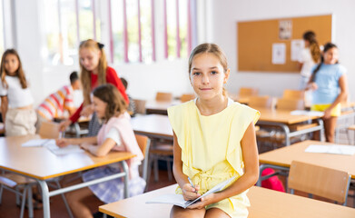 Happy tween schoolgirl repeating educational material before lesson, sitting during break on table with notebook in hands
