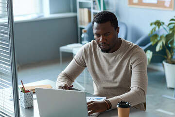Portrait of focused black man using laptop in office behind glass partition wall, copy space