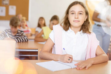 Portrait of interested diligent preteen schoolgirl sitting at school desk and writing exercises at lessons with classmates