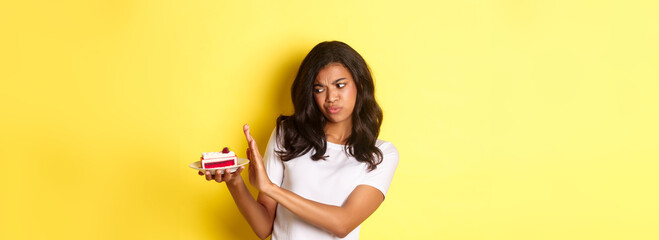 Image of displeased african-american girl decline to eat disgusting cake, standing over yellow...
