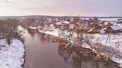 Winter river landscape, snow covered fields. Hoarfrost on trees, plants. Frosty evening. Sunny cloudy Misty weather. Cold season. Calm nature scene. Village