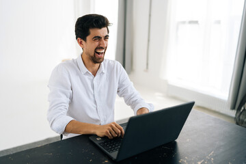 Studio portrait of happy excited young man in shirt using working on laptop computer smiling looking away sitting at table in room with light interior. Cheerful male freelancer at remote home office.