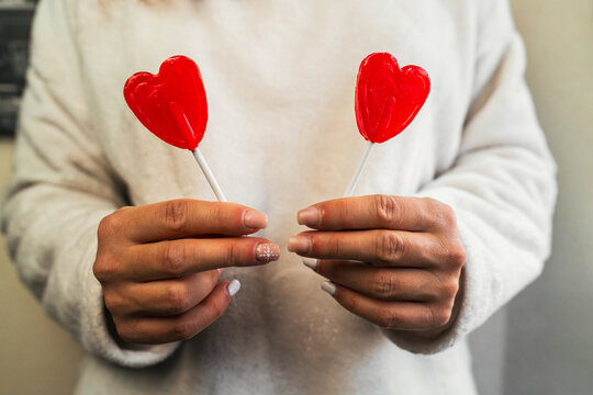 Close-up image of the hands of a woman wearing a white sweater holding two red heart-shaped lollipops in both hands. Young girl in love on Valentine's Day.