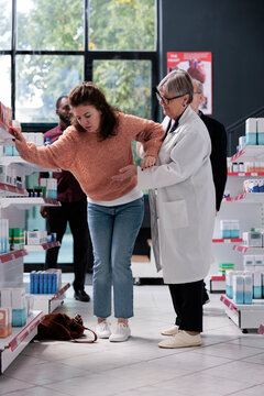 Woman Customer With Spinning Sensation And Dizziness Losing Balance While Being Helped By Senior Pharmacist Before Fainting In Pharmacy Shop. Health Care Service And Support