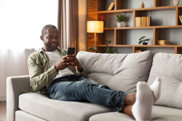 Smiling adult black guy using phone, chatting in social media, sitting on sofa in living room...