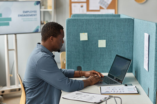 Side view portrait of black man working with laptop at workplace cubicle in office