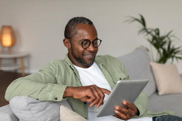 Happy adult black guy in glasses uses tablet for online work and business, sits on sofa in living...