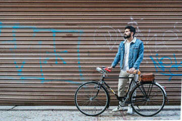 Young man in denim jacket holding his vintage classic bicycle in front of the closing shutter of a shop.