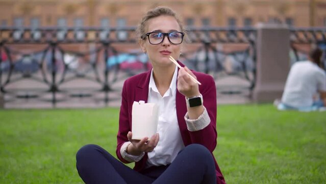 Young businesswoman having snack with takeaway food, sitting on lawn outdoors. Realtime