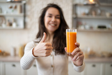Positive caucasian lady holding glass of organic orange juice and showing thumb up, standing in light kitchen interior