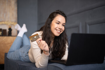 Excited young woman shopping online with credit card and laptop, lying on couch in living room...