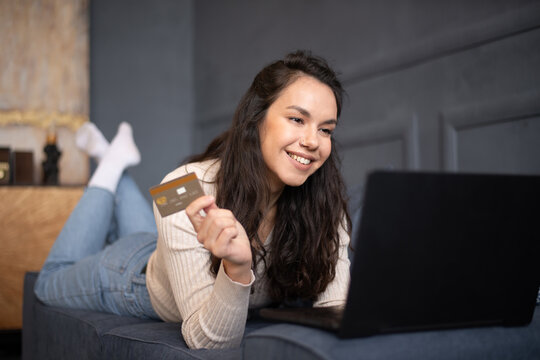 Excited young woman shopping online with credit card and laptop, lying on couch in living room interior, free space