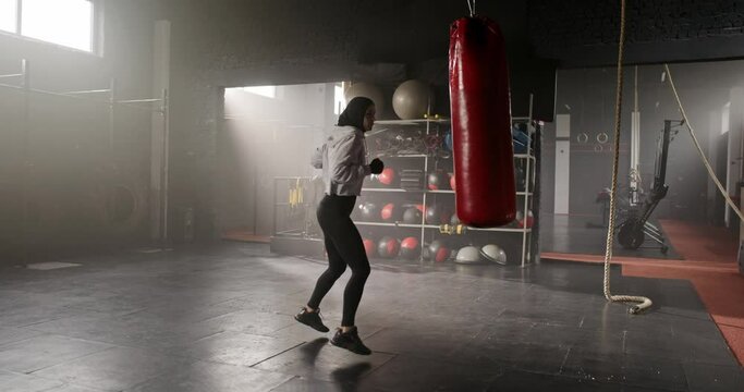 Side view of beautiful fit sporty Arab woman boxing fighter training with punching bag wearing hijab leggings and hoodie in crossfit gym. Attractive energetic muslim female working out boxing.