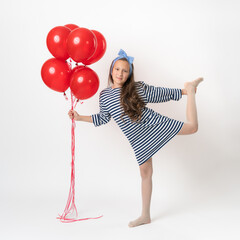Active girl posing, holding bunch of red balloons in hand and looking at camera. Caucasian model 10 years old in white blue striped dress standing on one leg on white background. Part of photo series