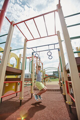 Vertical full length portrait of young boy playing on colorful playground in sunlight and hanging on monkey bars