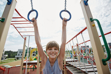 Front view portrait of smiling boy playing on colorful playground and hanging on monkey bars