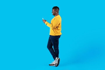 Fototapeta na wymiar Side View Of Young Black Male With Smartphone Standing Over Blue Background